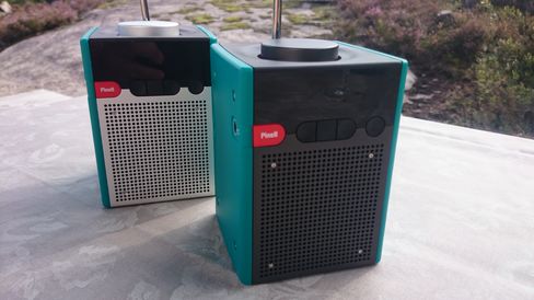 Den fremste radioen er en Pinell Go+, mens radioen i bakgrunnen er den originale Pinell-radioen uten Bluetooth. Den nye versjonen har mørke metallflater.
