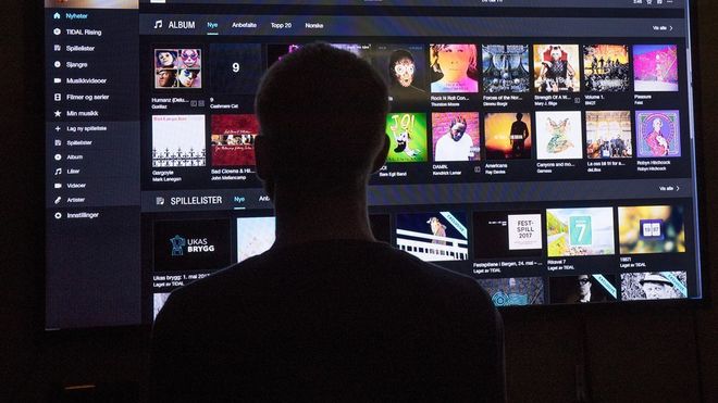 Silhuett av mann foran en stor skjerm som viser musikktjenesten Tidal og en rekke albumcovere.