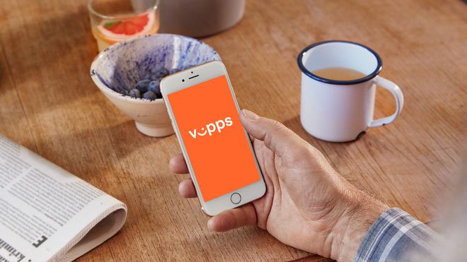 Vipps-logo på mobiltelefon i hånd. Kaffekopp og avis på bordet.