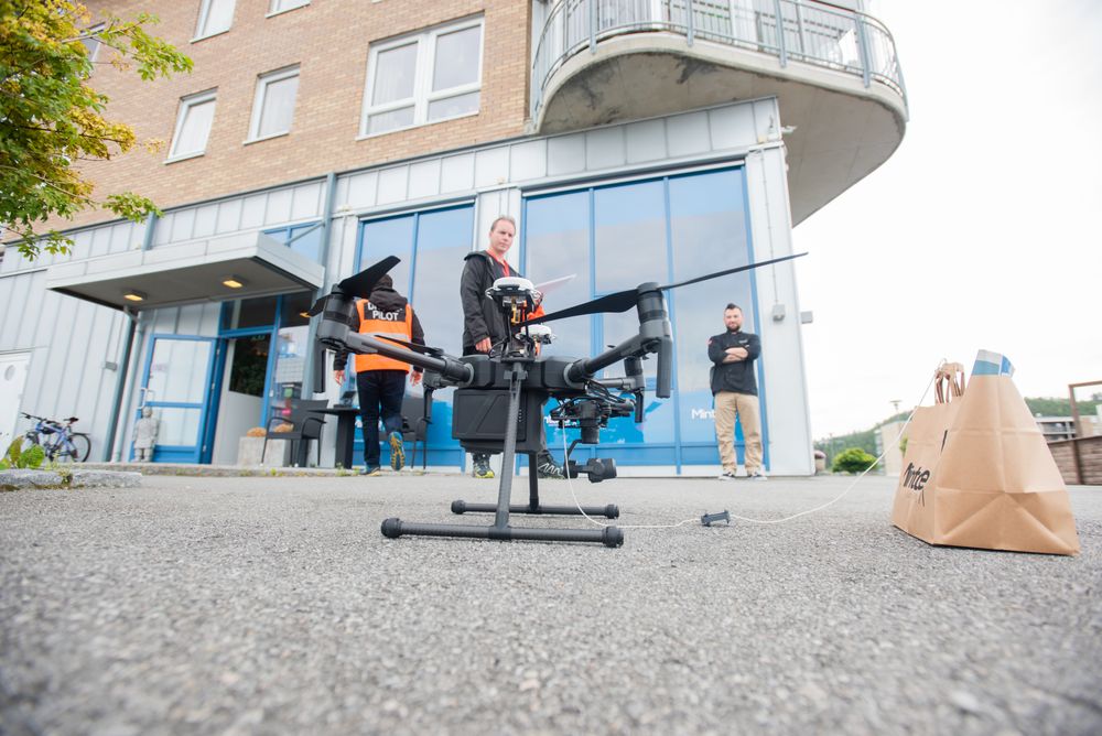 Posen med mat og avis er klargjort for flyfrakt med dronen. I bakgrunnen står Atilla Haugen (f.v.), Kristian Ystad og Kent Olav Ferstad.