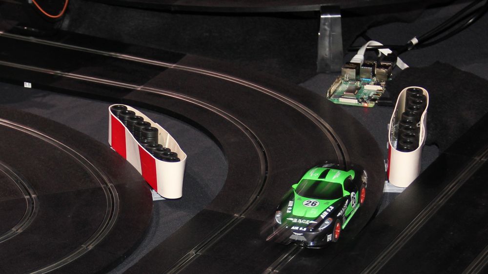 Racerbil i full fart ut av en sving på Oracles bilbane under JavaZone 2019. I bakgrunnen skimtes én av de mange små datamaskinene som brukes til å styre racerbilen autonomt.