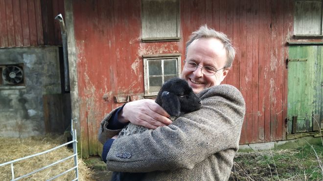 Håkon holder et sort lam foran en låve.