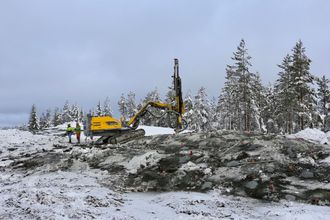To personer står ved siden av en borerigg på en høyde. Det er snø på bakken og du kan se furuskog i bakgrunnen.