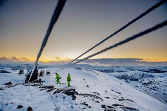 Tre kraftlinjer går inn i bildet der to personer arbeider på en fjelltopp. Det er dekket av snø og solen går ned i bakgrunnen.
