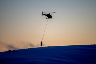 Et helikopter frakter en sylinder over en snøkledd fjelltopp i solnedgang.