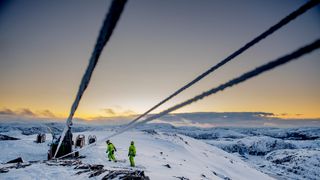 Tre kraftlinjer går inn i bildet der to personer arbeider på en fjelltopp. Det er dekket av snø og solen går ned i bakgrunnen.