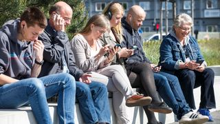 Seks mennesker, unge og gamle, sitter og ser ned i hver sin smarttelefon. 