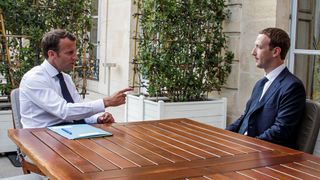 Emmanuel Macron og Mark Zuckerberg sitter ved et bord. Macron peker på Zuckerberg mens han ser ut til å gi en alvorspreken. 