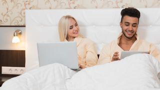 Ungt par sitter i en hotellseng og bruker henholdsvis en bærbar PC og et nettbrett.