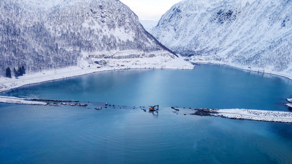 Gravemaskin «på fisketur» under arbeid på fyllingene på flo sjø sist vinter. Kryssingen av Sifjord på Senja skjer helt inne i fjordbunnen, for å unngå en rasfarlig veibit på sørsiden av fjorden.     