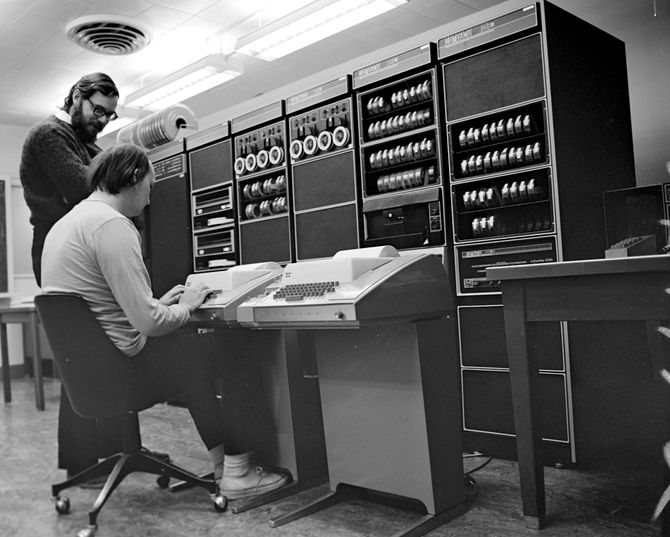 De to sentrale Unix-oppfinnerne, Dennis Ritchie (stående) og Ken Thompson, foran minidatamaskinen DEC PDP-11 og kabinetter med en hel del annet utstyr. Bildet er trolig tatt i 1972.