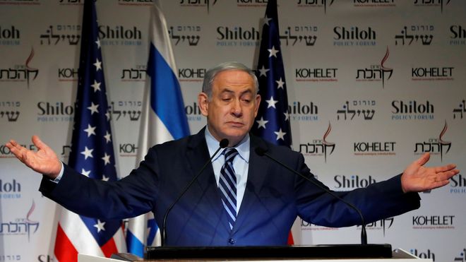 Benjamin Netanyahu slår ut med armene ved en taler stol foran amerikanske og israelske flagg. 
