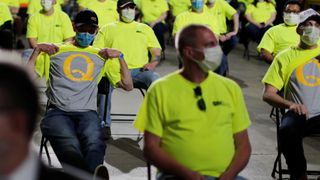 Mange ansatte i gule uniforms-t-skjorter sitter med ansiktsmasker. To av dem løfter opp de gule t-skjortene for å avsløre en skjorte med en Q på under.