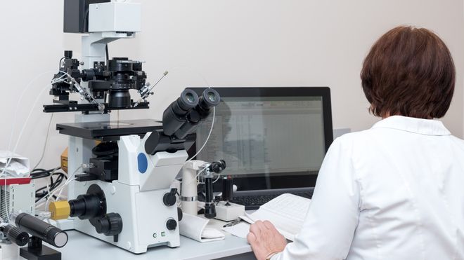 EN kvinne i hvit frakk sitter med ryggen til oss foran et mikroskop og en pc