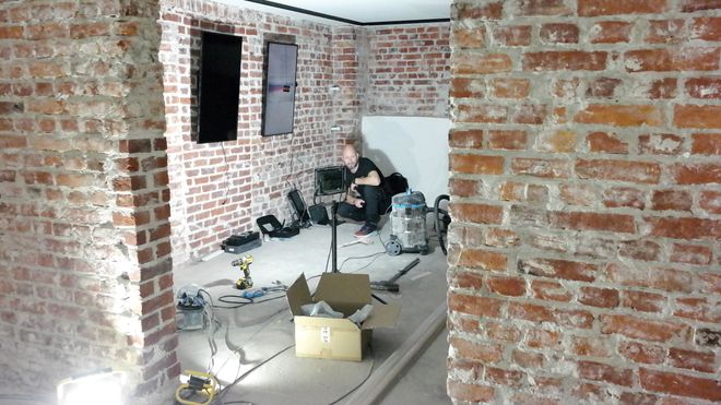 Et rom med bare murvegger, en arbeidslampe, litt rot og en stor støvsuger, en mann monterer skjermer på veggen