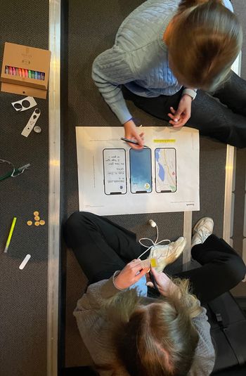 De to jentene sitter i en trapp med fargestifter og diverse og tegner og beskriver innholdet i mobiltelefoner printet ut på ark.