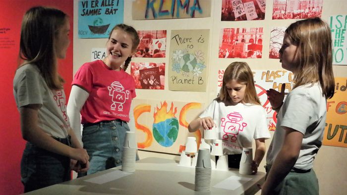 Tre jenter og en ung kvinne i rosa t-skjorte står i samtale rundt et bord med papirkopper-