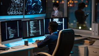 En mann sitter ved en pult med flere skjermer med kode foran seg og hendene på tastaturet.