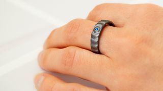 En kvinnehånd med en ring på langfingeren, grå med en blå stein og bølger i metallet.