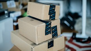 Tre pappesker med Amazon-logoer står på et bord i et rotete rom.