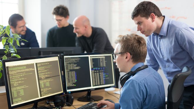En ung mann sitter ved datamaskinen og programmerer mens en annen står og ser på.