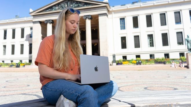En kvinne sitter på en benk på Universitetsplassen med en Macbook Air M2 i fanget