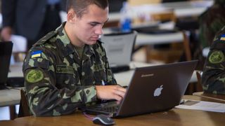 En soldat i med Ukrainas flagg på skulderen sitter foran en laptop.