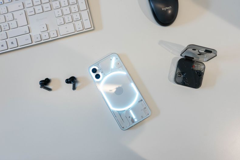 Nothing telefon på pult med Nothing øreproppene liggende til venstre, og dekslet til øreproppene til høyre, øverst i bildet ser man også et utsnitt av et tastatur og en mus.