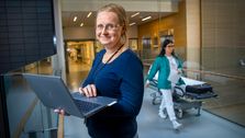 Tonje Holm Hjertaas står i en sykehuskorridor med en PC mens en sykepleier går forbi.
